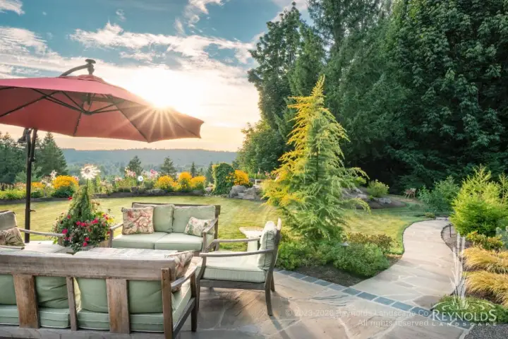 Back yard paver patio with outdoor lounge seating, market umbrella, and paver walkway bordered by lush garden beds and evergreens with sunset views