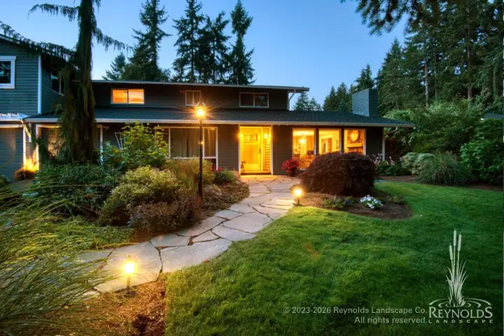 Random flagstone walkway with low-voltage path lighting leading to a mid-century home entrance surrounded by lush lawn and mature shrub borders at dusk