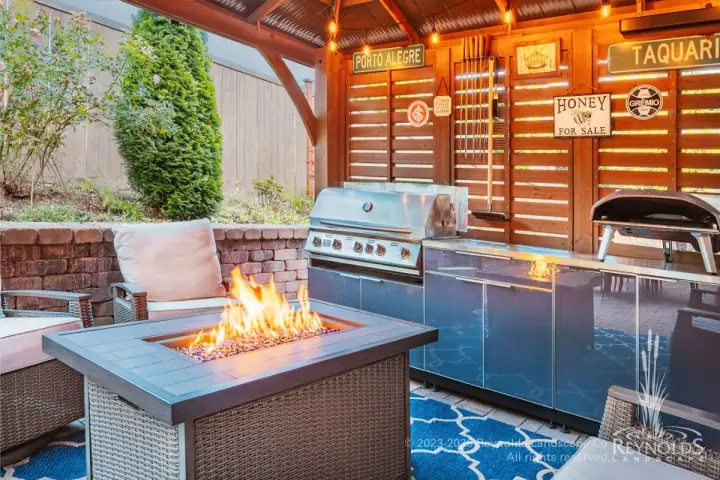 Rectangular gas fire table with glass media on covered patio with built-in outdoor kitchen, stainless steel grill, and wicker seating
