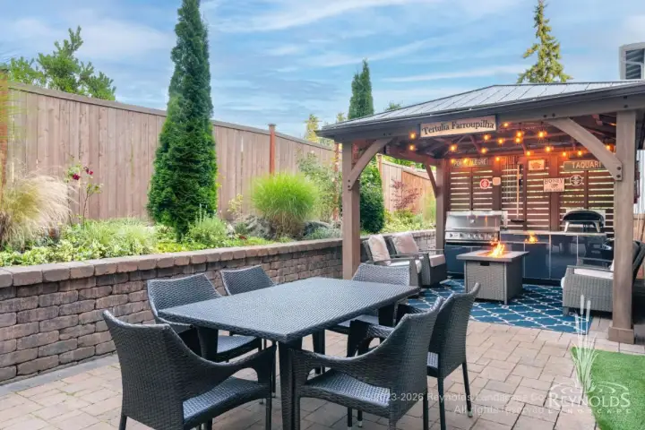 Paver patio with wicker dining set, pergola with string lights, and tall privacy hedge backdrop