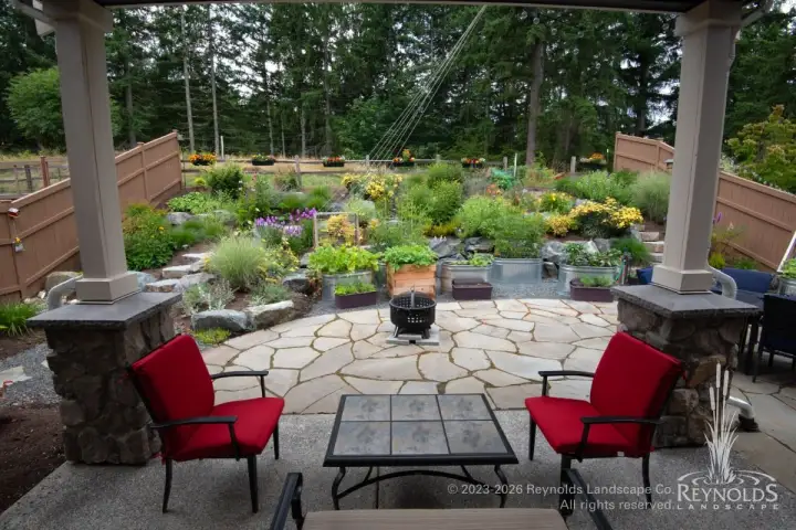 Back yard paver patio with fire pit, red lounge chairs, stone columns, and terraced garden beds with naturalistic plantings