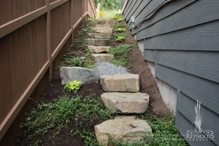 Narrow side yard with natural stone slab steps, ground cover plantings, and wood fence between homes