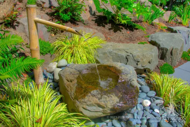 Naturalistic boulder pond with bamboo water spout surrounded by ornamental grasses, river rock, and lush garden plantings with hillside rockery backdrop