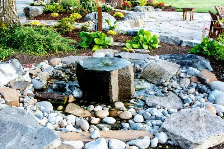 Bubbling urn water feature surrounded by river rock and decorative stone with garden border and paver patio backdrop