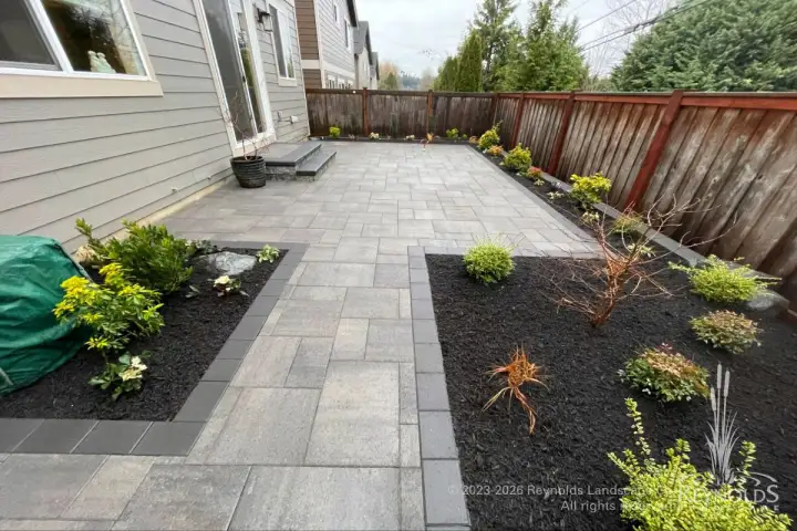 Back yard large-format paver patio with black mulch planting beds, young mixed shrubs, and wood privacy fence