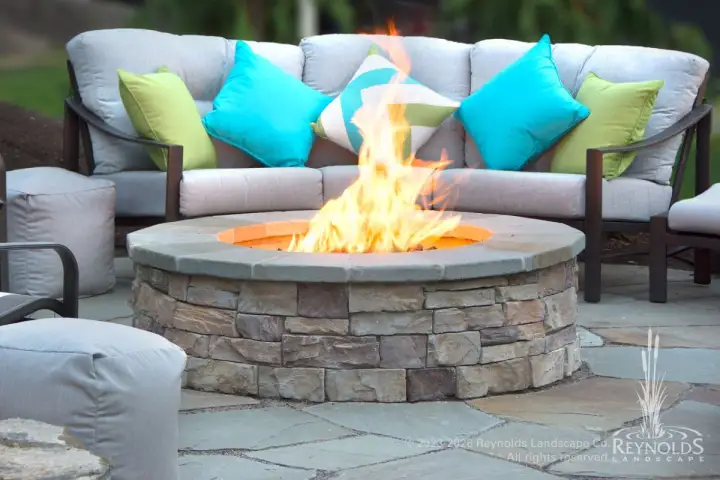 Round gas fire pit with stacked stone base and stone cap on patio with cushioned sectional sofa and colorful throw pillows