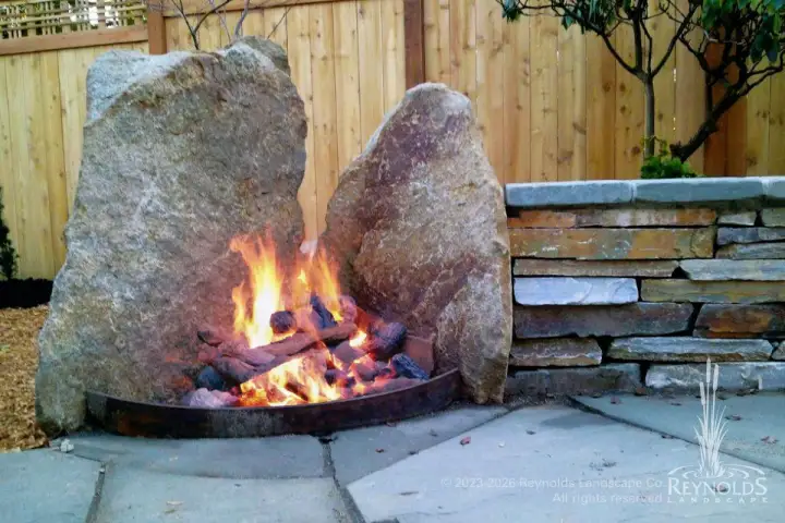 Two-boulder natural stone wood fireplace with steel basin on flagstone patio next to stacked stone retaining wall and wood fence