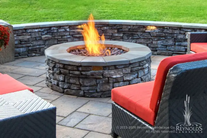 Round gas fire pit with stacked stone base and stone cap on paver patio with red cushioned wicker chairs and stone retaining wall