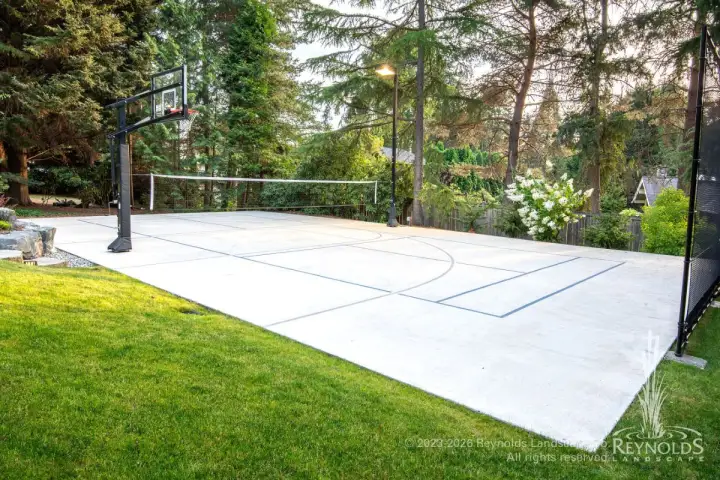 Multi-sport concrete court with basketball and volleyball net beside boulder retaining wall and garden