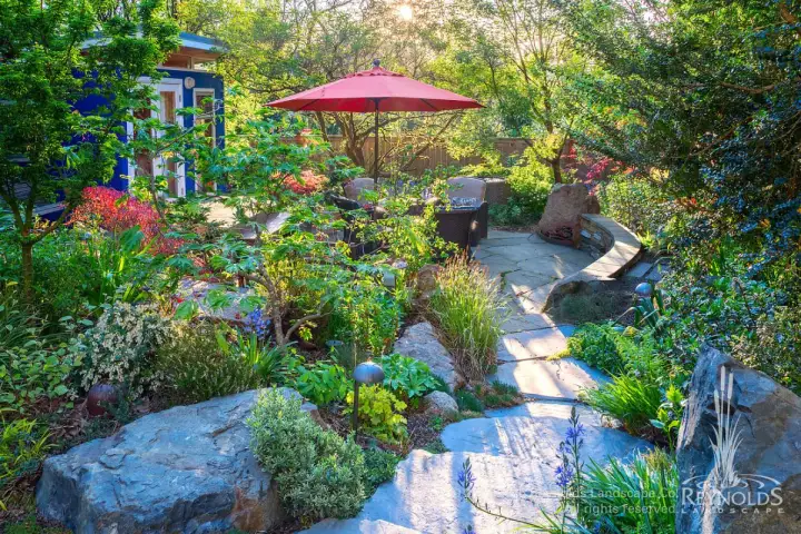 Lush cottage garden with flagstone paths, mixed perennial plantings, boulders, and a patio umbrella seating area