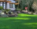 Sloped backyard lawn with stone slab steps, boulder retaining features, and mixed shrub plantings beside a modern home