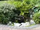 Small pond with boulder surround and river rock edging nestled among Japanese forest grass, dwarf conifers, and mixed evergreen shrubs in a Pacific Northwest garden