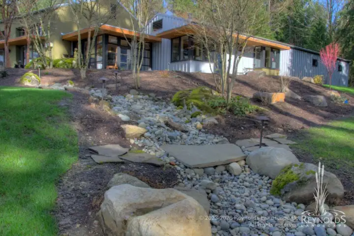 Dry river rock drainage swale with large boulders winding through sloped hillside landscape alongside single-story home