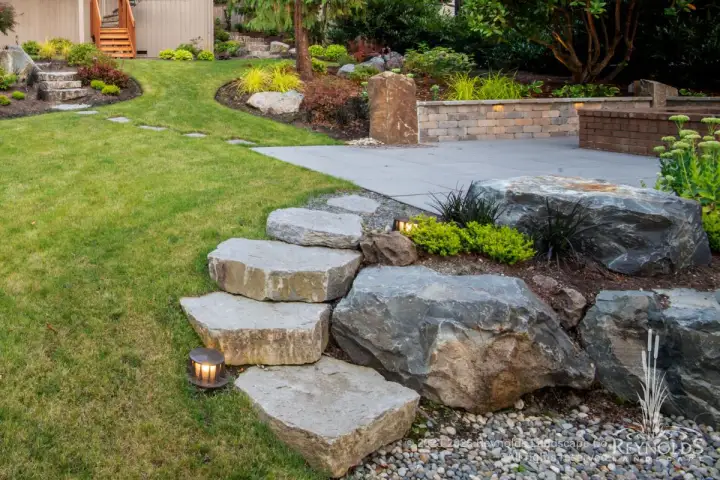 Natural stone slab steps alongside a large boulder leading to a concrete patio with lawn and garden beds