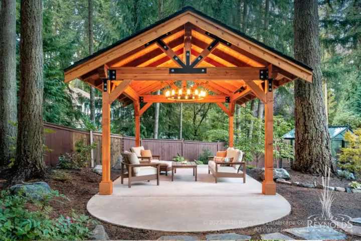 Freestanding timber frame pavilion with open gabled roof and chandelier over a cozy outdoor seating area surrounded by mature Pacific Northwest woodland