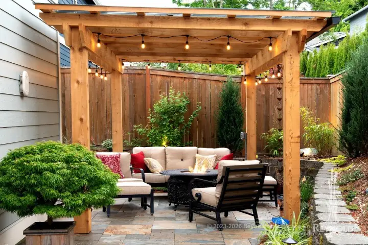 Cedar pergola with string lights over an outdoor lounge with wicker seating on a paver patio surrounded by mixed garden beds and a wood privacy fence