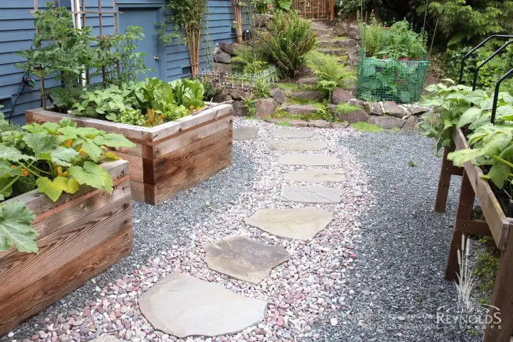 Flagstone stepping stone path set in river rock and gravel between cedar raised vegetable garden beds with fern-covered hillside steps in the background