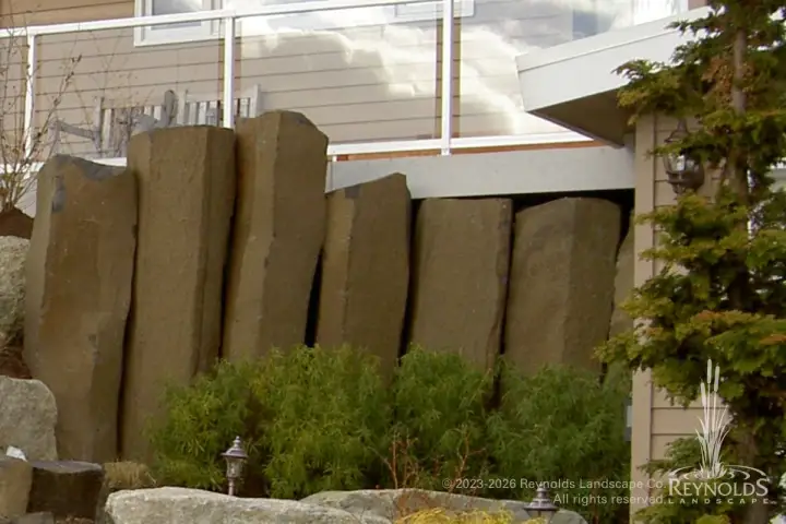Tall vertical basalt column retaining wall with ornamental plantings at base alongside modern home exterior