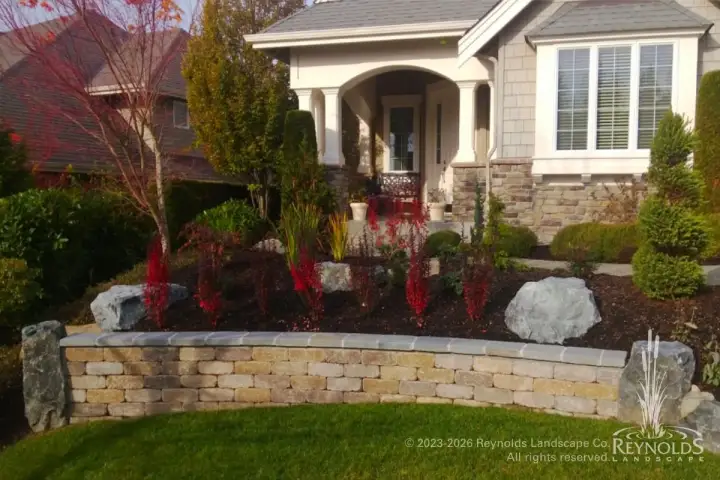 Curved concrete block raised planting bed with red flowering shrubs and ornamental plantings in front yard alongside craftsman home entry