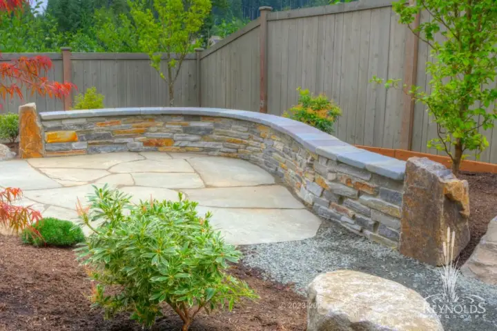 Natural stone seat wall with flagstone cap on flagstone patio with river rock border and ornamental shrubs alongside wood fence