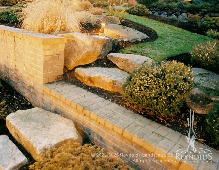 Tiered brick retaining walls with large flagstone steps, ornamental ground cover plantings, and manicured lawn backdrop