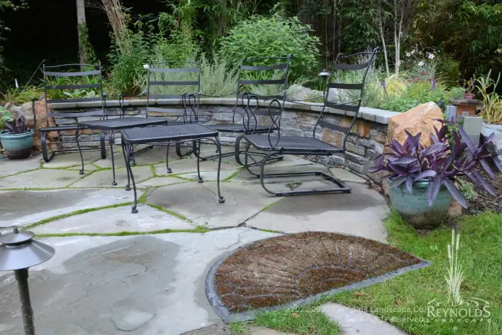Curved low stone seat walls with metal bench seating surrounding flagstone patio with planting bed and lush garden backdrop